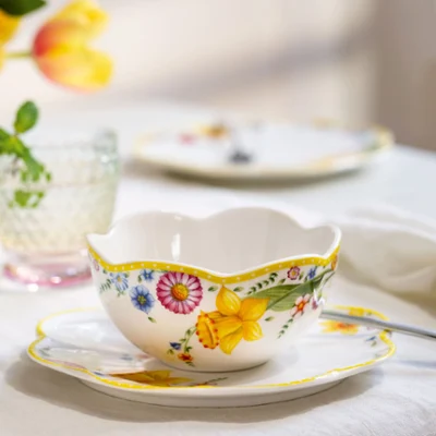 The ‘Spring Awakening’ teacup and saucer with spoon from Villeroy & Boch. The ‘Spring Awakening’ teacup and saucer with spoon from Villeroy & Boch stand on the table next to crystal glasses and softly blurred flowers in the background.