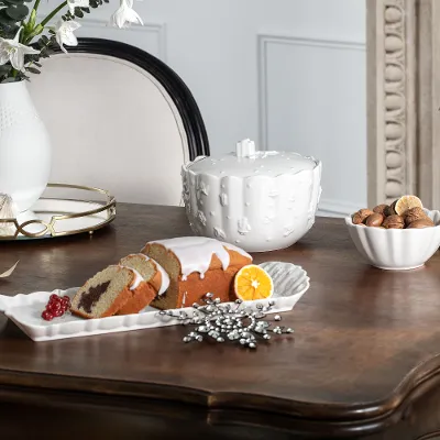 A wooden table with a cut cake on a Toy’s Delight Royal Classic platter from Villeroy & Boch, a bowl with nuts, a white decorative glass, a vase with flowers, candles, and Christmas decorations.