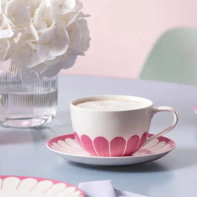A coffee-filled Fleur cup with pink leaf pattern on a Fleur saucer stands on a table with a Fleur vase with white flowers and matching tableware.
