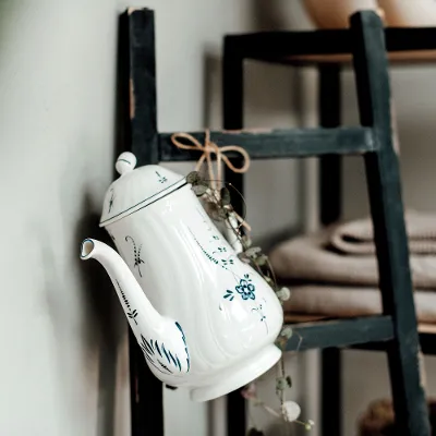 A white and blue Old Luxembourg teapot with a floral pattern hanging next to folded towels on a black wooden ladder.