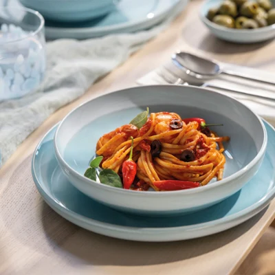 A pasta bowl stands on a blue Perlemor aqua plate. A pasta bowl from the Perlemor Aqua collection stands on a blue Perlemor Aqua plate with spaghetti, tomato sauce, and vegetables, next to cutlery and a salad.