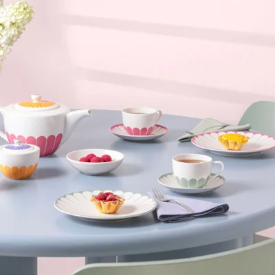A pastel-colored tea set with cups, saucers, plates with colorful floral patterns from the Fleur collection, and small desserts on a light blue round table.