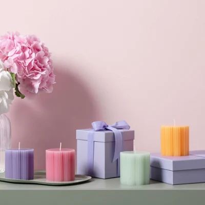 Velas Fleur en colores pastel como idea para regalo Jarrón de cristal Fleur con flores blancas y rosas, velas Fleur en colores pastel con forma de trébol y cajas de regalo moradas sobre una superficie verde delante de una pared rosa.