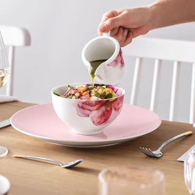 A person pouring dressing over a salad in a Rose Garden bowl at an elegantly laid dining table with Rose Garden crockery.