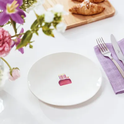 A With Love plate with an illustration of a cake on a table, with a fork and knife on a purple napkin. Next to this is a vase of pink and white flowers and a wooden board with croissants.