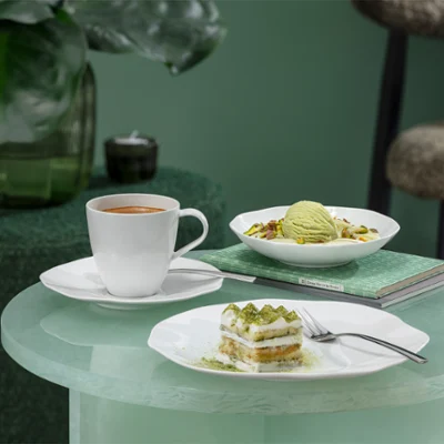 Eine Pura Kaffeetasse , ein Stück Kuchen und eine Pura Dessertschale stehen auf einem Glastisch Eine Pura Kaffeetasse , ein Stück Kuchen und eine Pura Dessertschale mit Eis stehen auf einem Glastisch mit einer Grünpflanze im Hintergrund.