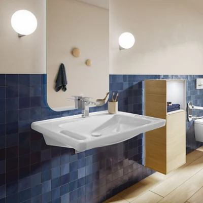 Un moderno cuarto de baño accesible con un lavabo rectangular ViCare con asideros para mayor seguridad, paredes de azulejos azules, un gran espejo, dos luminarias y una estantería de madera.