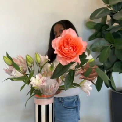 Memphis vase floral arranging Person partially hidden behind a lush coral flower arrangement in a striped vase, with pink lilies and leafy plants creating a warm, serene indoor scene.