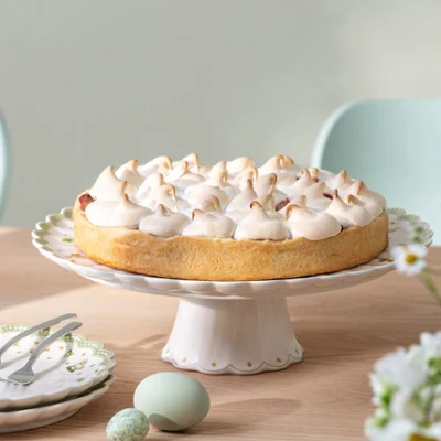 A cake with meringue stands on a wooden table on a cake stand with Easter decorations from Easter Delight, next to plates and flowers.