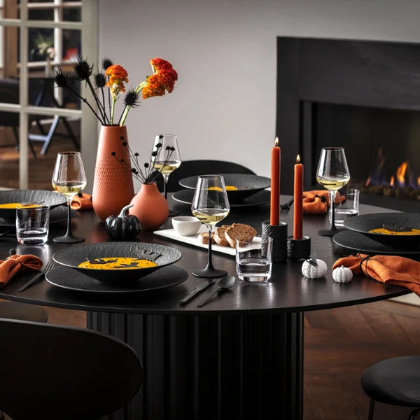 Una moderna mesa de comedor para cinco comensales con vajilla negra Manufacture, servilletas naranjas y decoración otoñal, incluyendo jarrones con flores y velas encendidas, junto a una chimenea encendida.