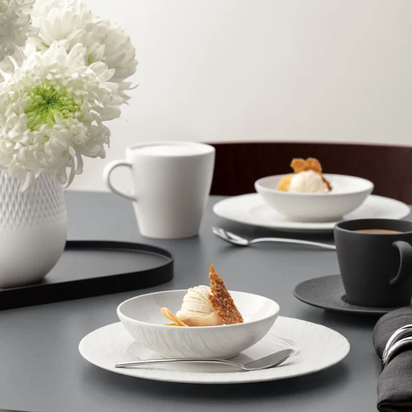 A formal table setting with a white bowl on a white plate with food on it, cup, and silver cutlery on a grey table.
