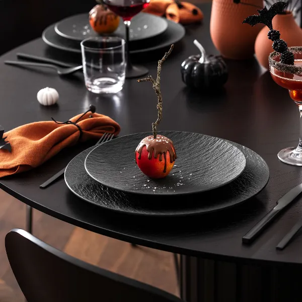 A black dining table is set with black Manufacture plates, orange napkins, and Halloween-themed drinks and decorations, including a chocolate-covered apple.