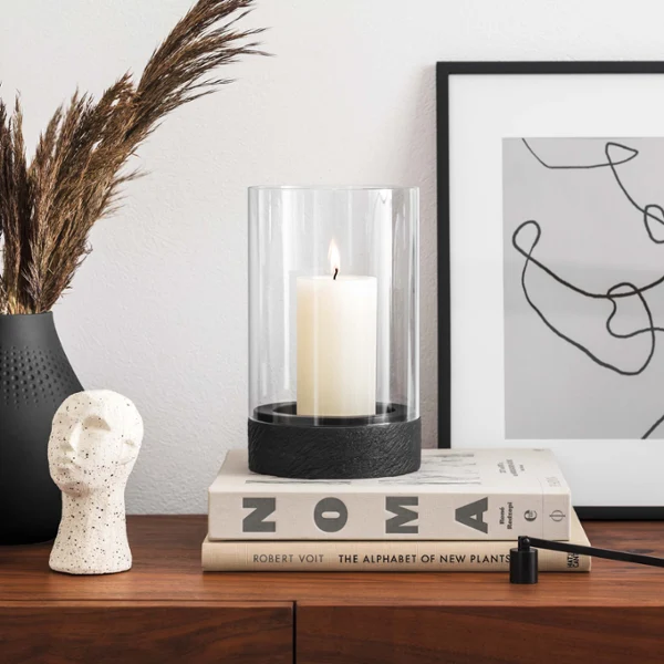 Świecznik dekoracyjny Manufacture na nowoczesnej szafce typu highboard Nowoczesna szafka typu highboard z przedmiotami dekoracyjnymi, w tym wazonem z suszonymi roślinami, czarnym świecznikiem dekoracyjnym Manufacture, rzeźbą, ułożonymi w stos książkami i oprawionym abstrakcyjnym dziełem sztuki.