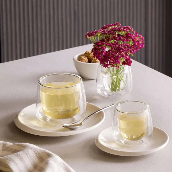 Una piccola teiera e una tazza di vetro di Villeroy & Boch NewMoon si trovano su un piatto bianco su un tavolo accanto a un vaso di vetro con fiori viola e una piccola ciotola con cubetti di zucchero di canna.