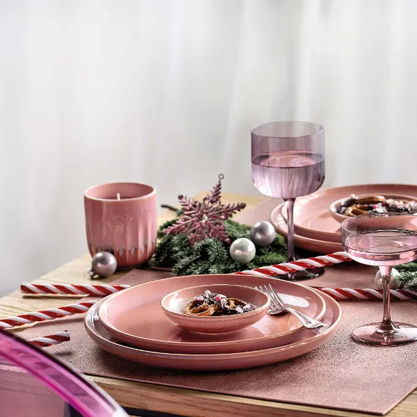 A festive table setting with pink Perlemor crockery, decorated with a snowflake ornament, candy canes, pink flowers and silver baubles. There are also two glasses, a pink candle and a small bowl of food.