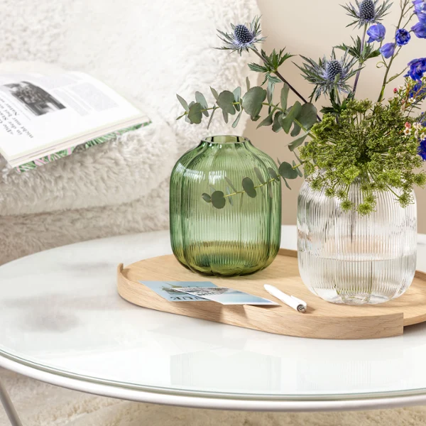 A round coffee table with a Villeroy & Boch Fleur vase holding blue and green flowers. In the background, an open magazine lies on a white, fluffy chair.
