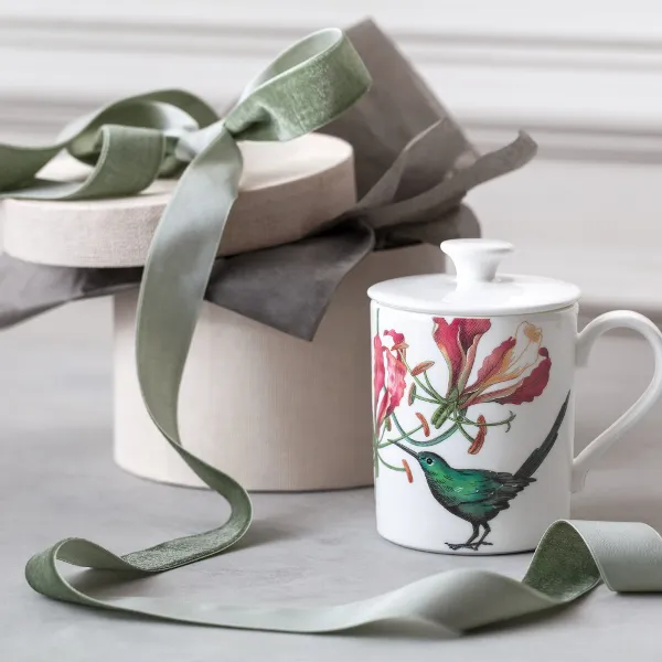 An Avarua mug from Villeroy & Boch with a bird and flower design next to a closed gift box decorated with a green bow on a light background.