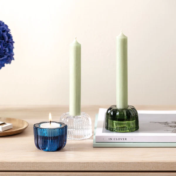 On a wooden table stands a Fleur vase and Fleur tealight holder from Villeroy & Boch, a blue flower arrangement adorns the vase, a burning candle in a blue holder, two green candles in holders, a book and matches.