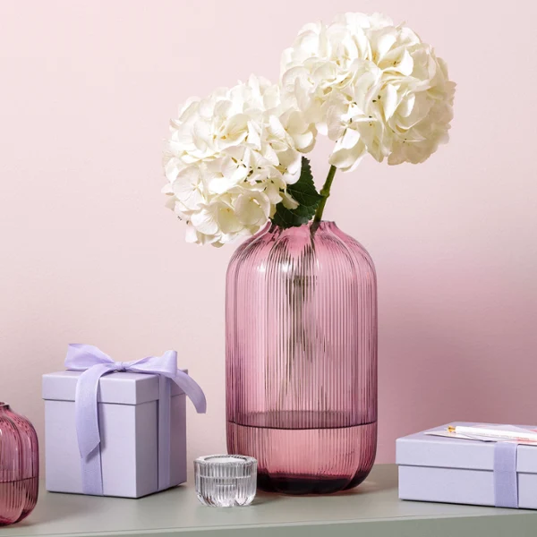 Pink glass vase by Fleur decorated with flowers A pink glass vase by Fleur with white hydrangeas next to two small lavender-colored gift boxes on a green surface.