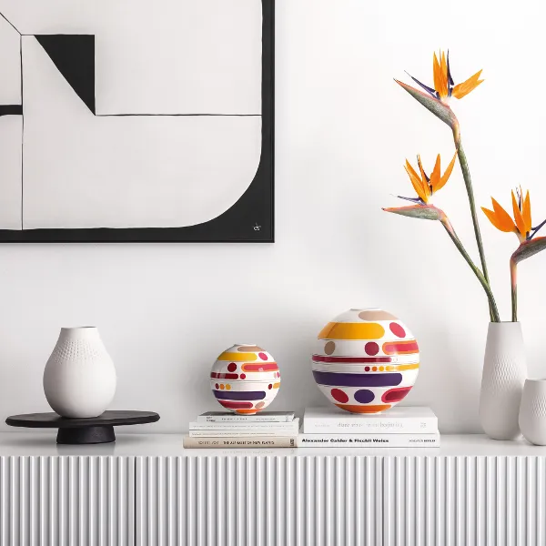 Una credenza minimalista espone la moderna La Boule Miami, vasi, fiori di uccello del paradiso e libri impilati sotto un'arte murale astratta.