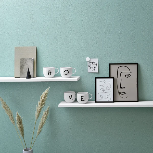Kubek Letters z literami H, O, M i E na półce Minimalistyczne półki z dekoracyjnymi przedmiotami, w tym kubki z napisem „Home", oprawiony w ramę abstrakcyjny obraz twarzy i roślina doniczkowa.