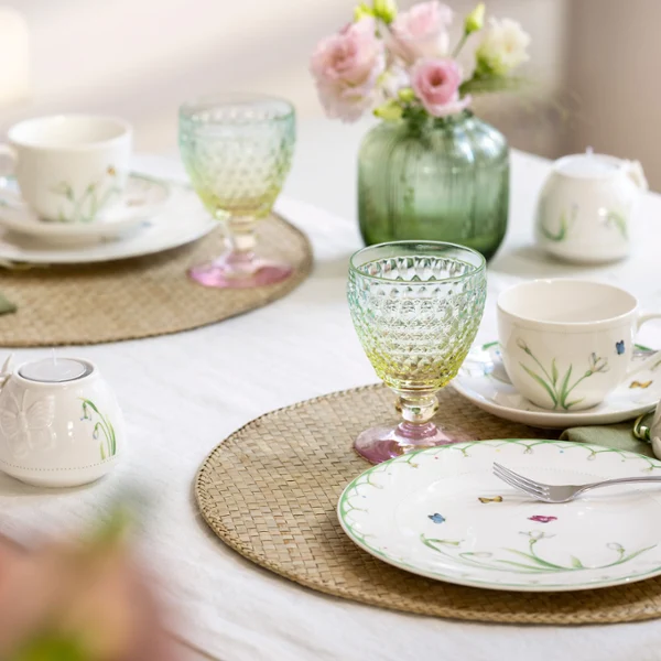 The Colourful Spring set from Villeroy & Boch embellishes any table for two with plates and cups with a floral pattern, bright green and yellow glasses, pink flowers in a vase and beige placemats on a white tablecloth.