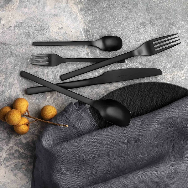Set di posate Manufacture nere con tovagliolo in tessuto e bacche decorative su una superficie grigia strutturata.