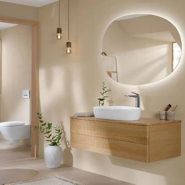 Antao bathroom with Oak wooden bathroom furniture and white ceramics in the typical teardrop shape A modern bright bathroom with a round backlit mirror above a floating wooden washstand with a white washbasin. Two pendant lights hang from the ceiling. There is a potted plant on the floor next to the floating washbasin.