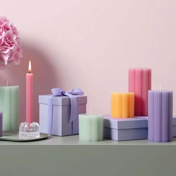 Świece Fleur w różnych pastelowych kolorach i kształtach jako idealny pomysł na prezent Różne pastelowe świece Fleur i ozdobne pudełka na prezenty na zielonym tle z różowymi i białymi kwiatami w tle.