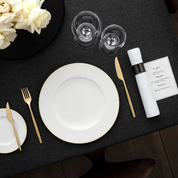 Une table noire dressée avec de la vaisselle blanche raffinée Château Septfontaines Une table dressée avec des assiettes blanches Château Septfontaines, des couverts de table dorés, deux verres, une serviette roulée et une composition florale sur une nappe foncée.