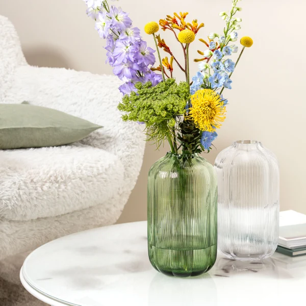 A round coffee table with a Villeroy & Boch Fleur vase holding blue and green flowers. In the background, an open magazine lies on a white, fluffy chair.