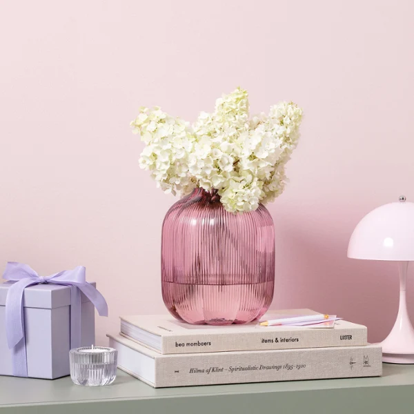Vase en verre rose de Fleur avec des fleurs blanches sur des livres empilés, à côté d'un coffret cadeau violet et d'une petite lampe de table rose.