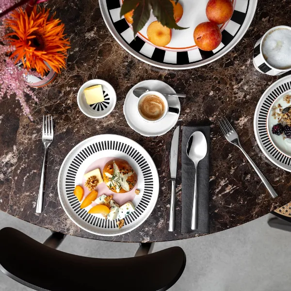Vista dall'alto di un tavolo di marmo con ciotole di frutta, caffè, cereali e yogurt della collezione Memphis di Villeroy & Boch, posate e decorazioni floreali.