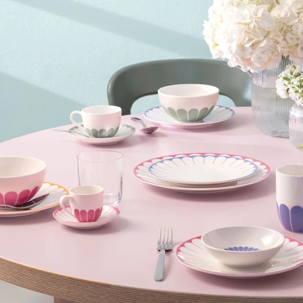 Okrągły stół nakryty zastawą Villeroy & Boch Fleur Okrągły stół z talerzami, kubkami, filiżankami, sztućcami, szklankami i białymi wazonami z kwiatami od Villeroy & Boch Fleur.