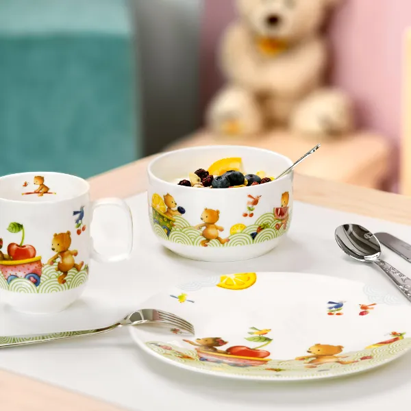 De la vaisselle pour enfants Happy as a bear avec des fruits et des biscuits Un ensemble pour le petit-déjeuner Hungry as a bear avec un bol de céréales et des fruits sur une table, ainsi qu’une tasse, des couverts de table et un ours en peluche en arrière-plan.