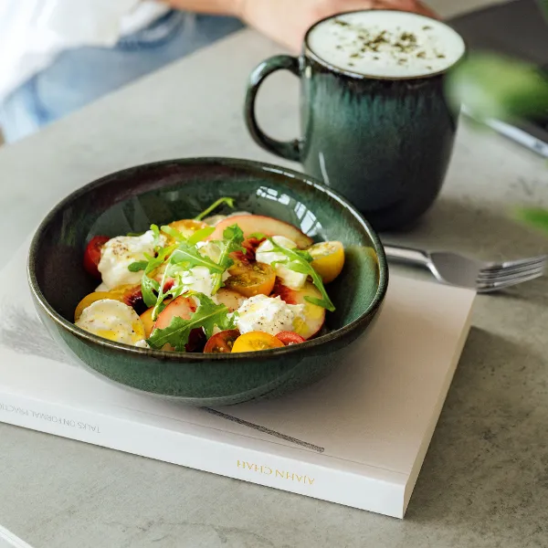 A salad with tomatoes, lettuce, and cheese is presented on a book in a green Lave bowl from Villeroy & Boch, accompanied by a frothy drink in a matching green mug on a grey background.