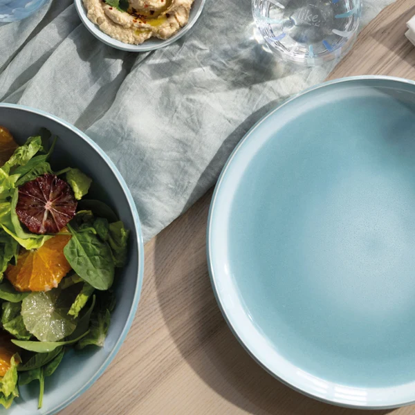 An empty blue Perlemor Aqua plate stands next to a bowl of salad. An empty blue Perlemor Aqua plate stands next to a bowl of salad, cutlery and a napkin on a wooden table.