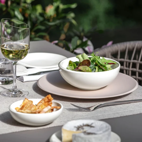 White NewMoon bowl with salad presented on a beige dinner plate A Villeroy & Boch NewMoon bowl filled with salad beside wine glasses.