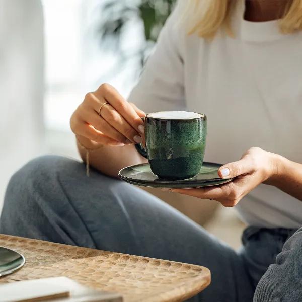 A person in a white shirt and jeans sits on a sofa and holds a green Lave mug from Villeroy & Boch with a frothy drink in his hand. In the foreground is a wooden table with a plate of toast.