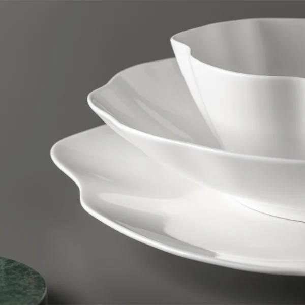 Drei weiße Pura Teller mit gezacktem Rand sind auf einer grauen Fläche Drei weiße Pura Teller mit gezacktem Rand sind auf einer grauen Fläche neben einem runden, grünen Objekt gestapelt.