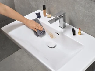 A hand is cleaning a washbasin in the bathroom with a grey cloth and removing a make-up stain. There are various make-up products on the rim of the washbasin.