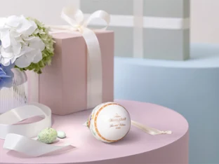 A pastel pink gift box with a white bow sits on a pink table next to the white and gold Annual Easter Ornament, with a bow, light green sweets and a vase with white and blue flowers. A blue gift box stands in the background.