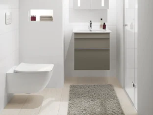Modernes Badezimmer mit weißen Wänden, schwebendem Venticello-Waschbecken mit Spiegelschrank, wandmontiertem WC und verglaster Dusche. Handtücher und Toilettenartikel sind ordentlich in einem vertieften Regal angeordnet. Beiger Teppich auf dem Boden.