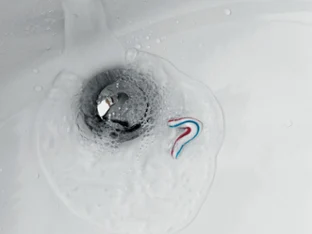 A small amount of toothpaste and water is flowing down the drain of a bathroom washbasin, causing bubbles to form around the drain cover.