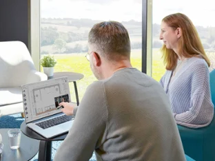 Two people are sitting in front of a laptop. The man is pointing at the screen.