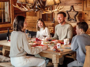 A family of four sits at a wooden table in a rustic room, eating together and chatting.