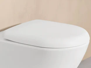 A close-up of a white closed toilet lid in front of a light beige wall.