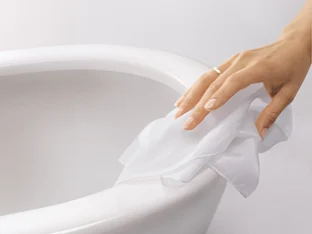 A hand wearing a ring wipes a white cloth over a white ceramic toilet.