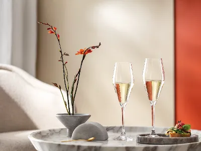 La Divina Gläser mit Sekt gefüllt Ein Paar La Divina-Gläser von Villeroy & Boch stehen auf einem Marmortisch, begleitet von orangefarbenen Blumen in einer Vase und einem Canapé, und sorgen für ein elegantes Ambiente im stilvollen Wohnzimmer.
