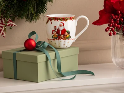 Annual Chistmas edition 2026 mug A green gift box with a bow, a red Christmas ornament, and a mug with a Santa Claus motif from Villeroy & Boch in front of a white background, with Christmas decorations visible in the background.